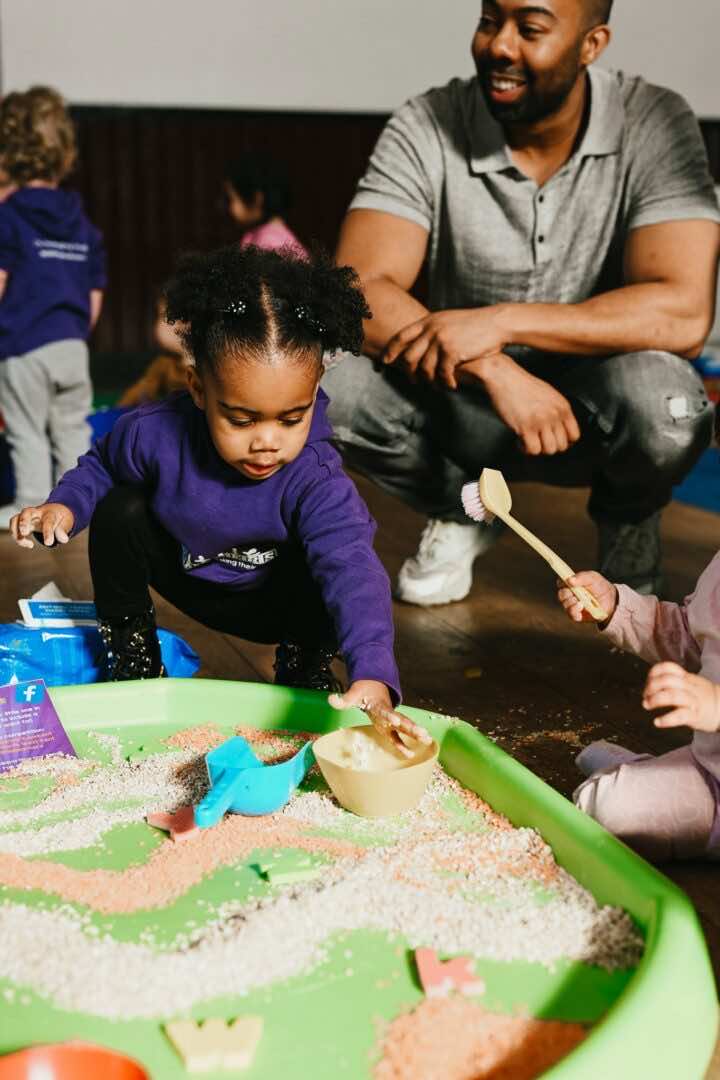 Daddy watches on as their child plays in a tuff tray at a Little Learner's Messy Play Class